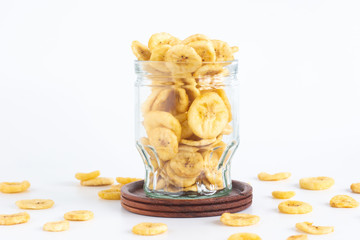 Dried banana slices in glass bottle with wooden saucer isolated on white background