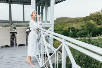 Relax time. Beautiful young woman wearing at white robe stay on luxury terrace with cup of coffee. Vacation in mountains.