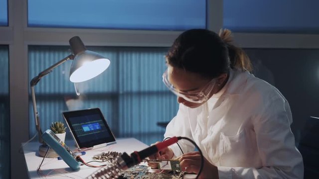 Close up of female engineer of electronics in white coat and protective glasses checking motherboard with multimeter tester and other electronic devises