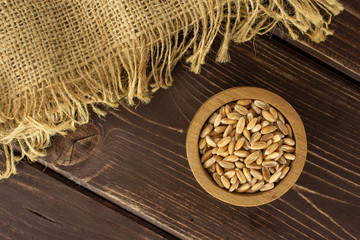 Lot of whole fresh beige dinkel wheat grain in a wooden bowl and jute cloth flatlay on brown wood