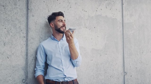 Young businessman leaning against wall and talking on phone with speaker mode