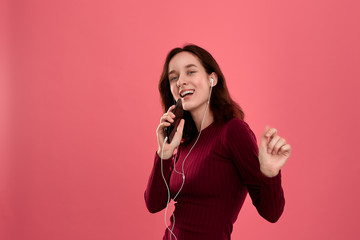 Happy young brunette woman singing and dancing using smartphone as a microphone and wearing wired headset.