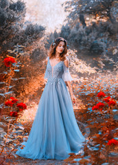 sad princess walks in fading autumn garden with withered plants, lady with short dark hair in chic light blue sky dress looks at red roses with sadness, clean young attractive girl, creative colors. © kharchenkoirina
