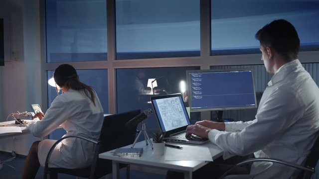 Team of electronics engineers in white coats working on the computers in advanced laboratory. Smart people working together. Back view