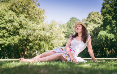 Naklejka na ściany i meble Summer portrait of young woman sitting on the grass, in sunny day. Beautiful, romantic woman looking to the sky.
