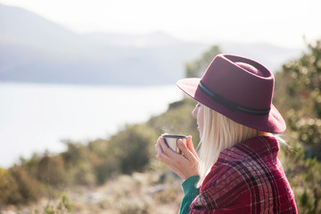Woman traveler drinks coffee in autumn mountains above sea. Cozy fall picnic on beach. Girl in warm...