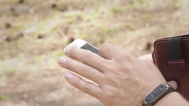 Male using smartphone outdoors in nature. Hands visible, fingers touching touchscreen. Browsing or texting context.