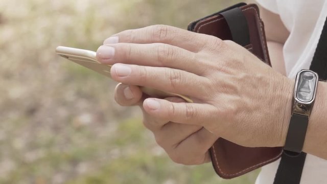 Male using smartphone outdoors in nature. Hands visible, fingers touching touchscreen. Browsing or texting context.