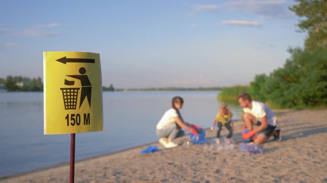 signpost on trash can, young family with little child collects refuse in garbage bag on dirty beach from plastic and polyethylene while cleaning near river in unfocused background