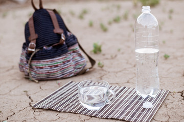 A cup of water and a walking backpack on the dried ground. The concept of thirst, dehydration, the need to drink water.