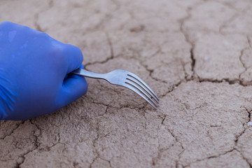 The concept of food shortages, environmental disaster. A rubber gloved hand holds a fork on the background of cracked dry ground.