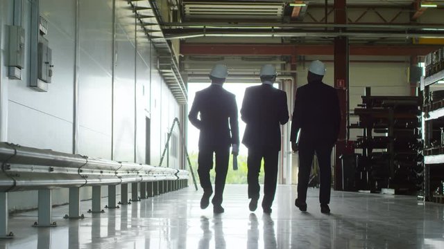 Pan wide backlit shot of three executives dressed in suits and white safety helmets departing production facility warehouse after inspection, strolling towards bright sunlit exit and talking
