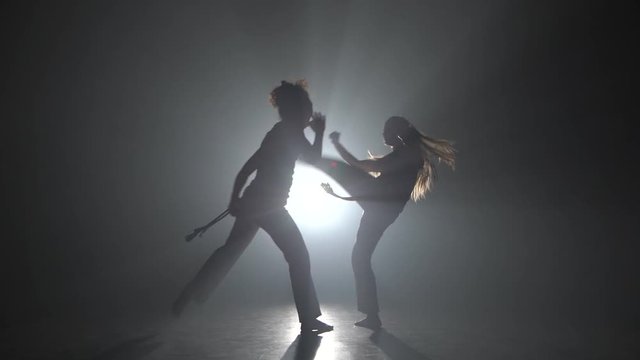 Strong ladies practicing capoeira in darkness against spotlight in studio.