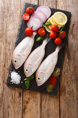 Uncooked dorado or gilt-head sea bream fish with spices, tomato, onion and lemon close up on a slate board. Vertical top view