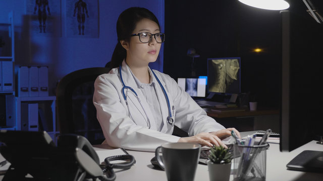 Asian Female Doctor In Dark Clinic Office Typing On Computer At Night. Young Woman Medical Staff Nurse Working Concentrated In Midnight Hospital Alone. Smart Lady With Eyeglasses Serious Face.