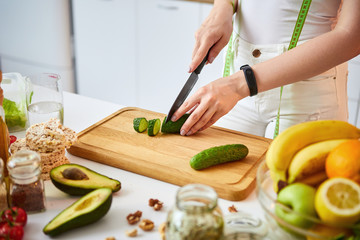 Young happy woman cutting cucumber for making salad in the beautiful kitchen with green fresh ingredients indoors. Healthy food and Dieting concept. Loosing Weight