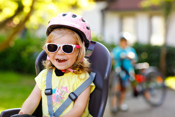 Portrait of little toddler girl with security helmet on the head sitting in bike seat of parents. Boy on bicycle on background. Safe and child protection concept. Family and weekend activity trip.