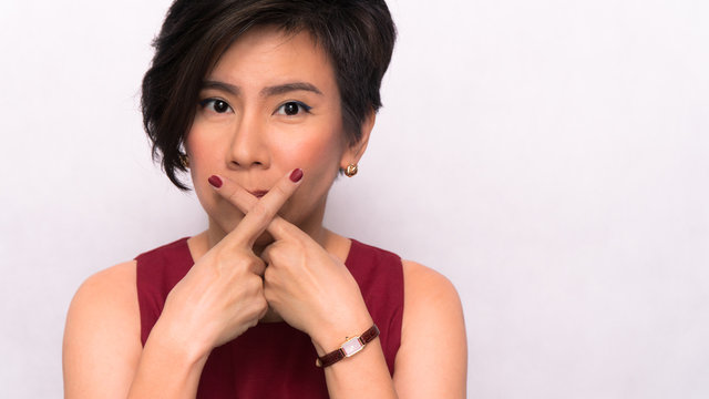 Studio portrait of a beautiful asian woman in stylish red outfit, holding fingers crossed at her lips, shows taboo sign. Keep secret, Quit smoking, Girl gossip, No bad word, Don't talk, Mute concept.