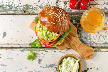 Vegan burger with carrot and avocado in classic bun, served on wooden board with avocado sauce and ingredients above on white wood texture background. Flat lay, space. Healthy fast food.