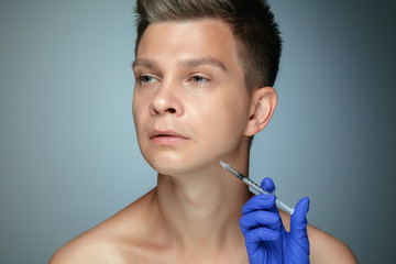 Close-up portrait of young man isolated on grey studio background. Filling surgery procedure, lips and cheekbones. Concept of men's health and beauty, cosmetology, body and skin care. Anti-aging.