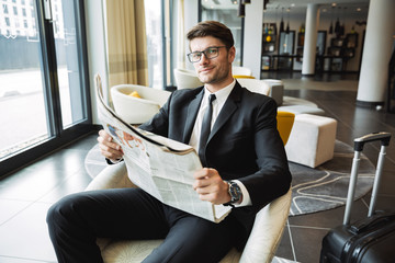 Portrait of pleased young businessman sitting on armchair and reading newspaper in hotel hall
