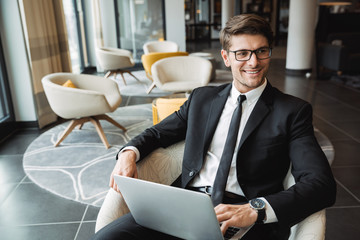 Portrait of satisfied young businessman sitting on armchair with laptop computer in hotel hall
