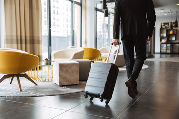 Cropped photo of caucasian businessman wearing suit walking with suitcase in hotel lobby