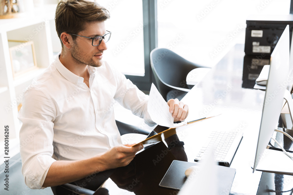 Poster image of masculine businesslike man holding clipboard with papers while working on computer in offic