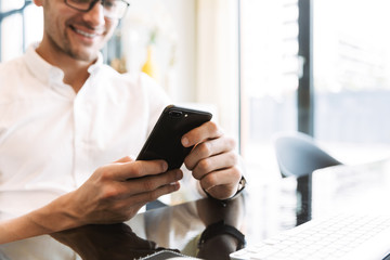 Image of smiling businesslike man holding cellphone while working on computer in office