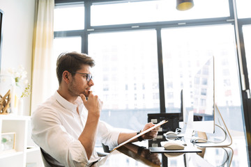 Image of confident businesslike man holding clipboard with papers while working on computer in office