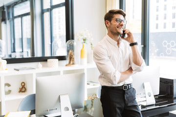 Image of joyful businesslike man talking on cellphone while working in office