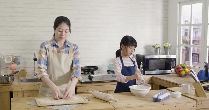 slow motion cute asian singing little girl wearing apron mixing ingredients in metal bowl on wooden kitchen background. elegant mom making dough beside. two parent and kid spend time handmade.