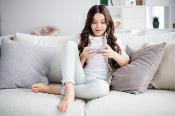 Portrait of her she nice attractive charming cute cheerful cheery focused wavy-haired pre-teen girl sitting on divan using internet online browsing in light white interior living-room house indoors