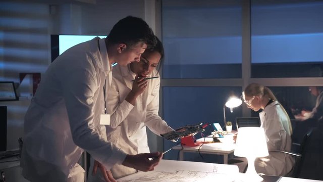 Technology Research Laboratory: Mixed race electronics engineers in white coats working with motherboard and control electronics scheme in lab. They discussing many questions