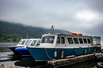 Fototapeta premium IOANNINA, GREECE - JUNE 6, 2019 - Three small old tourist cruise white and blue ferryboats moore on lake Pamvotida near the beautiful small Greek town. Early morning spring view with unusual fog