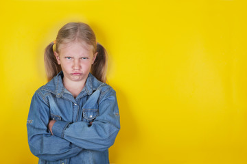 Little funny girl dressed in blue jeans shirt standing with arms folded on yellow background. ...
