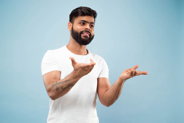 Half-length close up portrait of young hindoo man in white shirt on blue background. Human emotions, facial expression, ad concept. Negative space. Showing empty bar, pointing, choosing, inviting.
