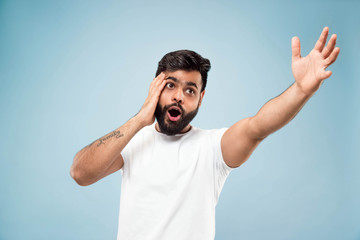 Fototapeta premium Half-length close up portrait of young hindoo man in white shirt on blue background. Human emotions, facial expression, sales, ad concept. Negative space. Pointing up being shocked and astonished.
