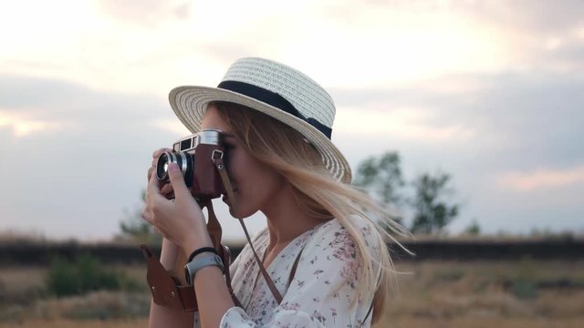 Young beautiful girl in white dress stands in field with vintage camera, on background sunset. Cheerful woman in wicker hat taking photos with retro camera on the meadow. Slow motion, close up.