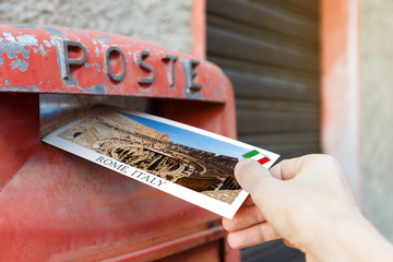 Hand drops a postcard in a red mailbox in the Rome, Italy. The postcard shows a panorama of...