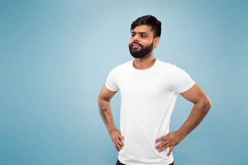 Half-length close up portrait of young hindoo man in white shirt on blue background. Human emotions, facial expression, ad concept. Negative space. Posing, standing and smiling, looks calm.
