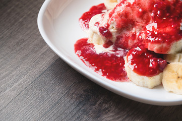 Closeup Vanilla ice cream poured with strawberry or raspberry jam with sliced banana slices on a white plate on a dark wooden table. You can do this at home on vacation