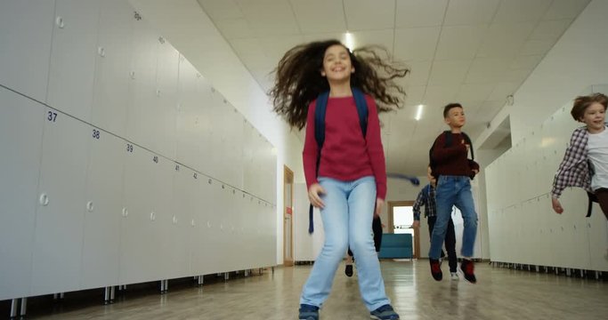 Cheerful Caucasian small teen schoolgirl with a schoolbag running and jumping at the school coridor after lessons or at the break with her school friends and classmates.