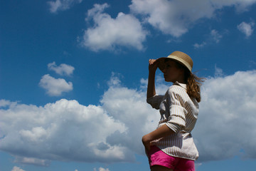 Cropped shot of a young girl, wearing a hat and white striped shirt, standing and looking to the side over blue sky background. Young girl outdoors. People, Travel, Nature concept.