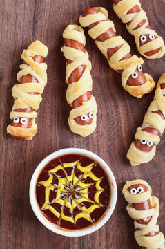 Fun food for kids. Mummy hot dogs lying on a rustic table with a bowl of ketchup and mustard dip, with spider web design. Top view, flay lay position.