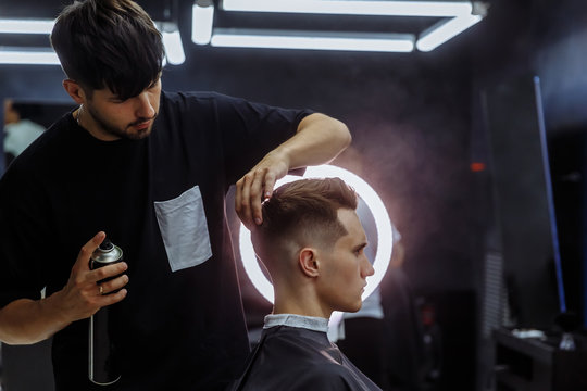 Barber makes hair styling with hair spray after haircut at the barber shop. Young handsome Caucasian man getting a haircut in a modern hairsalon.
