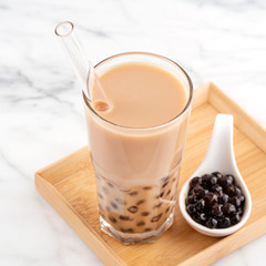 Tapioca pearl ball bubble milk tea, popular Taiwan drink, in drinking glass with straw on marble white table and wooden tray, close up, copy space.