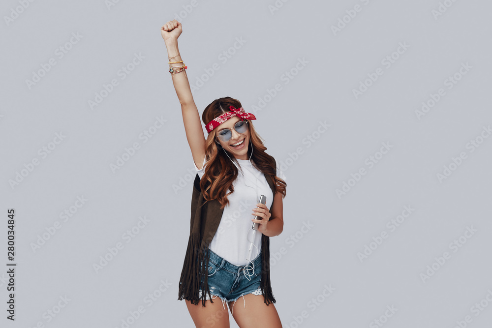 Poster Attractive stylish young woman listening music and smiling while standing against grey background