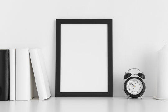 Black frame mockup with books, clock and a candle on a white table.Portrait orientation.