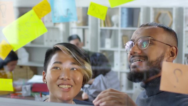 Close-up shot of two multiethnic team members standing in front of glass wall, discussing post-it reminders with tasks, pointing, talking and smiling, with colleagues sitting at table in background